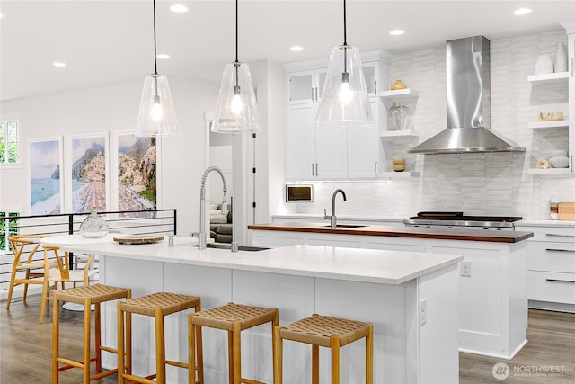 kitchen with a kitchen island with sink, a sink, wall chimney range hood, open shelves, and stainless steel gas stovetop