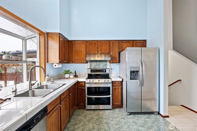 kitchen featuring brown cabinets, a sink, under cabinet range hood, tile countertops, and appliances with stainless steel finishes