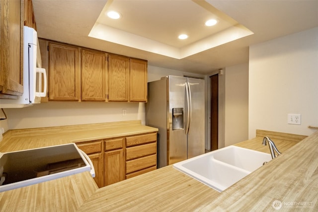 kitchen with recessed lighting, a raised ceiling, brown cabinetry, a sink, and stainless steel fridge with ice dispenser