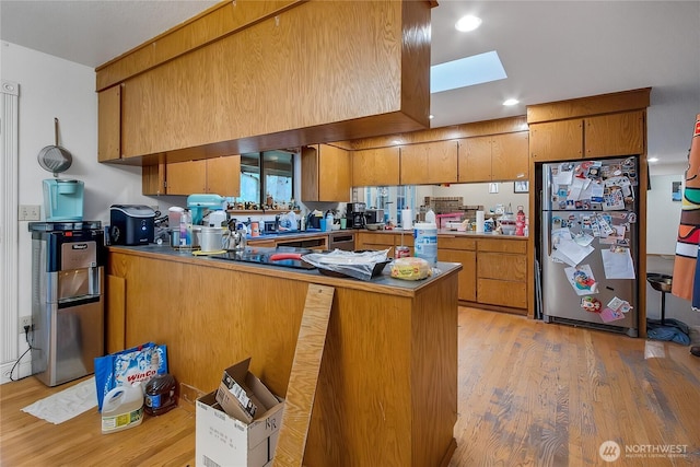 kitchen featuring a skylight, light wood-style flooring, brown cabinetry, freestanding refrigerator, and a peninsula