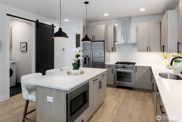 kitchen featuring a barn door, wall chimney exhaust hood, stainless steel appliances, a kitchen bar, and a sink