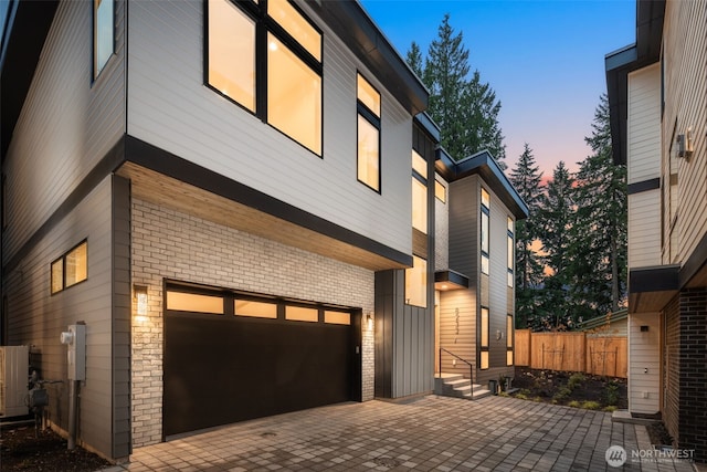 property exterior at dusk with an attached garage, fence, decorative driveway, and brick siding