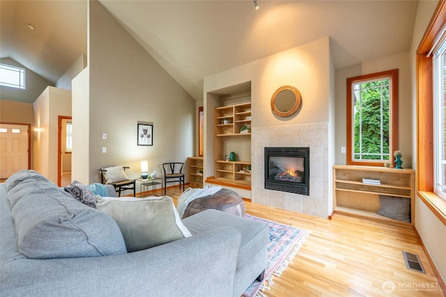 living area with high vaulted ceiling, a tile fireplace, wood finished floors, visible vents, and built in features