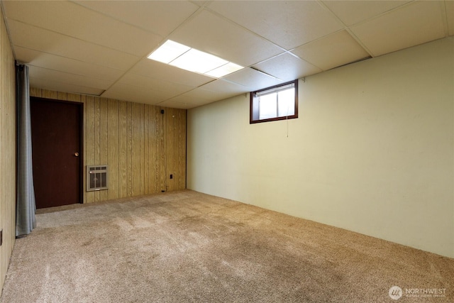 basement featuring a paneled ceiling, light colored carpet, wood walls, and heating unit