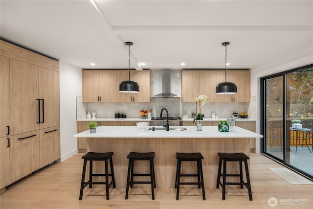 kitchen with wall chimney range hood, a center island with sink, light countertops, and decorative light fixtures