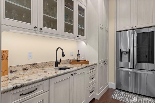 kitchen with a sink, white cabinets, light stone countertops, stainless steel fridge, and glass insert cabinets