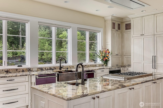 kitchen with white cabinets, an island with sink, glass insert cabinets, light stone countertops, and stainless steel gas stovetop