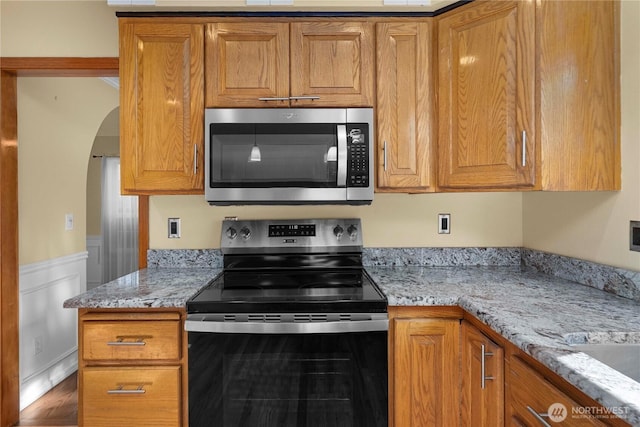 kitchen with light stone counters, brown cabinetry, arched walkways, and stainless steel appliances