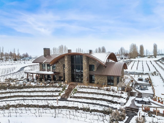 snow covered rear of property featuring a standing seam roof, stone siding, a chimney, and metal roof