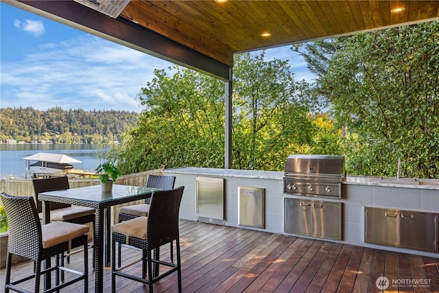 deck with area for grilling, a water view, a sink, a view of trees, and an outdoor kitchen