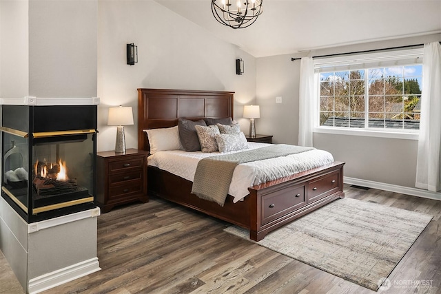 bedroom featuring lofted ceiling, a multi sided fireplace, wood finished floors, a chandelier, and baseboards