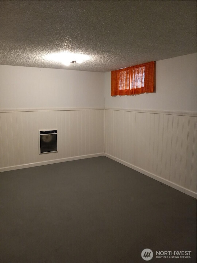 finished below grade area featuring dark colored carpet, wainscoting, a textured ceiling, and heating unit