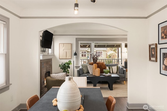 dining room with wood finished floors, baseboards, a fireplace, arched walkways, and ornamental molding