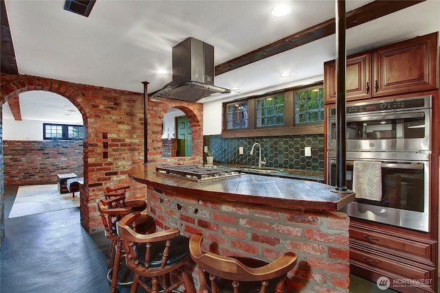 kitchen featuring island range hood, arched walkways, brick wall, stainless steel appliances, and a sink