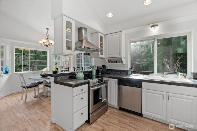 kitchen featuring lofted ceiling, stainless steel appliances, wall chimney range hood, white cabinetry, and a sink
