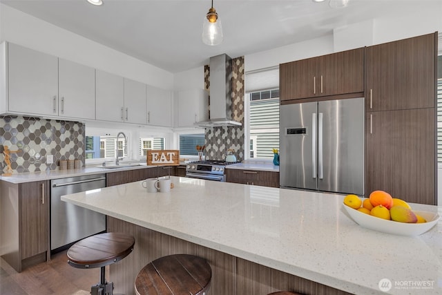 kitchen featuring decorative backsplash, wall chimney exhaust hood, appliances with stainless steel finishes, a kitchen bar, and a sink