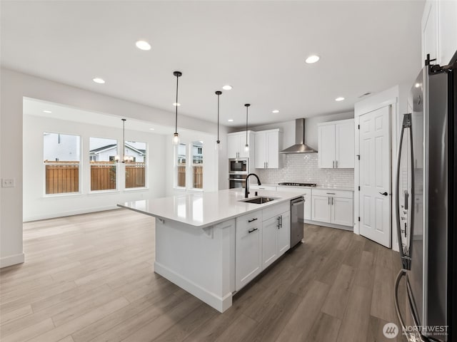 kitchen with stainless steel appliances, light countertops, white cabinets, wall chimney range hood, and an island with sink