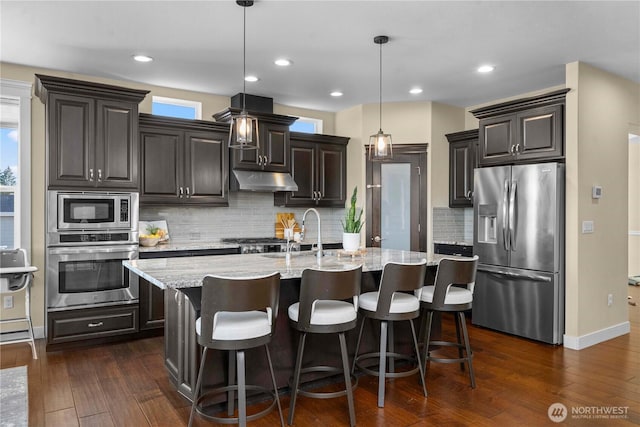 kitchen featuring light stone counters, dark wood-style flooring, a center island with sink, stainless steel appliances, and under cabinet range hood