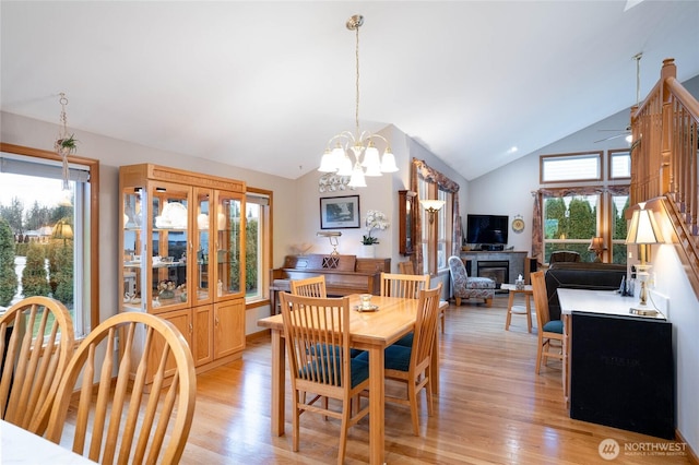 dining room featuring light wood finished floors, ceiling fan with notable chandelier, high vaulted ceiling, and a glass covered fireplace