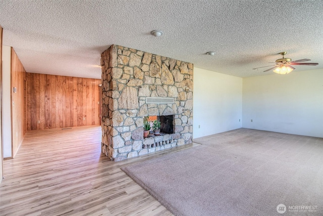 unfurnished living room featuring a textured ceiling, a stone fireplace, wooden walls, wood finished floors, and a ceiling fan