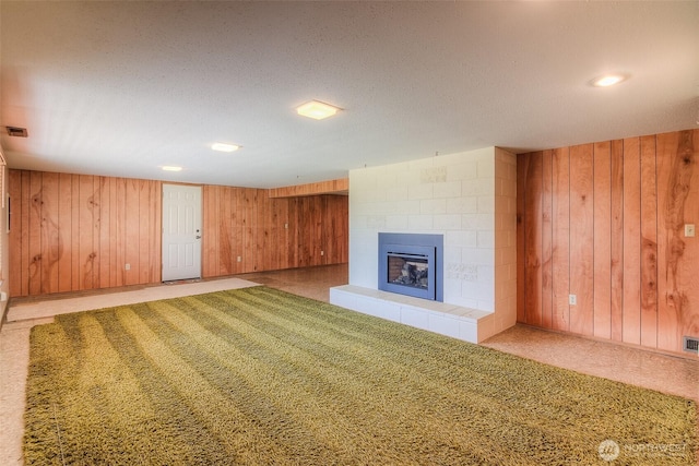 unfurnished living room featuring a textured ceiling, wood walls, carpet flooring, visible vents, and a tiled fireplace