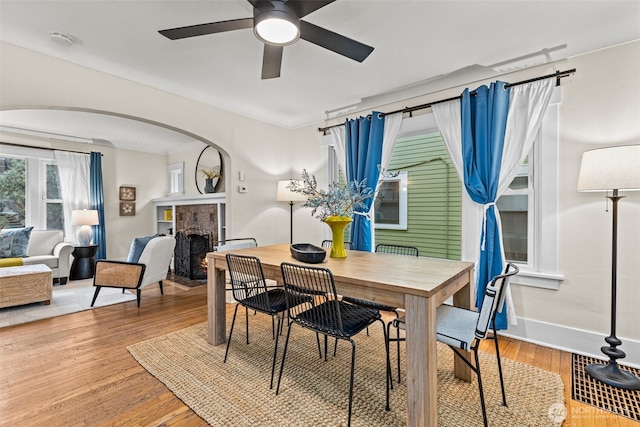 dining room with a warm lit fireplace, baseboards, arched walkways, a ceiling fan, and wood finished floors