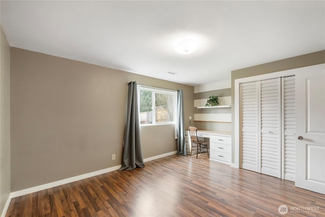 unfurnished bedroom featuring a closet, baseboards, built in desk, and dark wood-style flooring