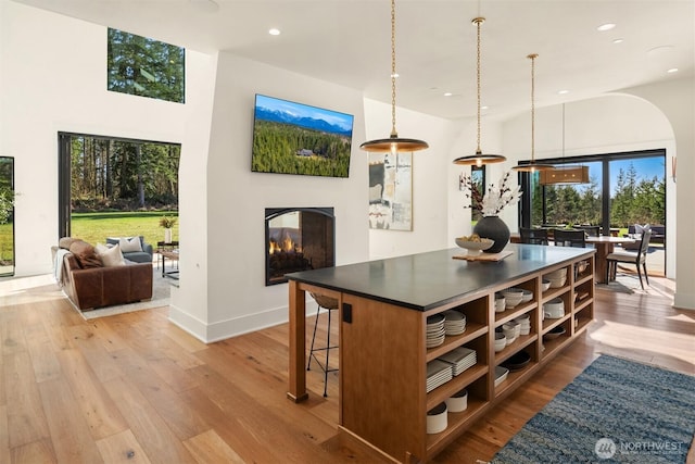 kitchen featuring a multi sided fireplace, light wood-type flooring, a center island, open shelves, and dark countertops