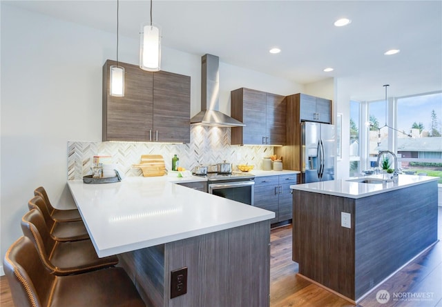 kitchen featuring pendant lighting, a breakfast bar area, light countertops, appliances with stainless steel finishes, and wall chimney exhaust hood