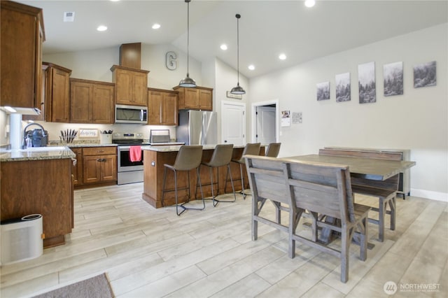 kitchen with brown cabinets, hanging light fixtures, appliances with stainless steel finishes, a kitchen island, and light stone countertops