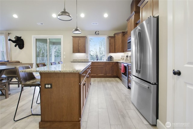 kitchen featuring a kitchen island, hanging light fixtures, appliances with stainless steel finishes, brown cabinetry, and a kitchen bar