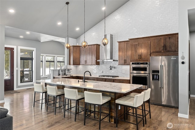 kitchen featuring wood finished floors, stainless steel appliances, wall chimney range hood, high vaulted ceiling, and a sink