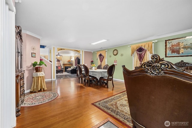 dining area featuring arched walkways, a fireplace, wood finished floors, baseboards, and crown molding