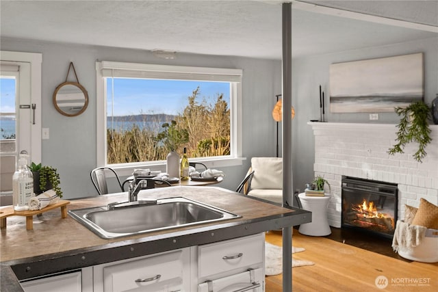 kitchen with a sink, white cabinets, a brick fireplace, light wood finished floors, and dark countertops