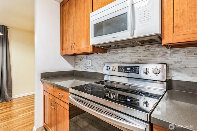 kitchen featuring tasteful backsplash, brown cabinetry, dark countertops, white microwave, and stainless steel range with electric cooktop