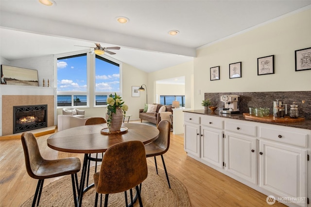 dining room featuring a ceiling fan, light wood finished floors, lofted ceiling, a lit fireplace, and a water view