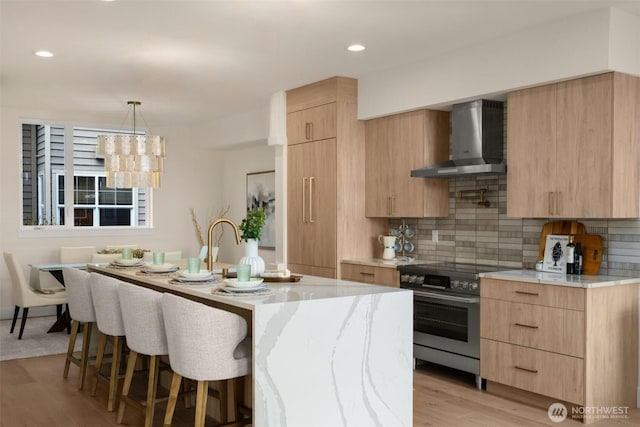 kitchen featuring electric range, a kitchen island with sink, light brown cabinetry, wall chimney range hood, and a kitchen bar