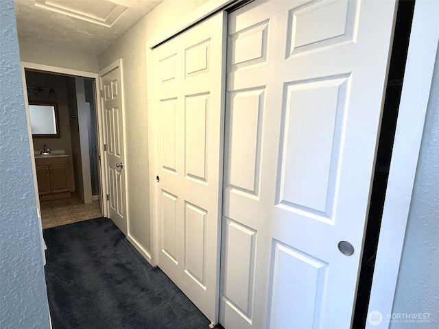 corridor featuring a sink, a textured wall, dark colored carpet, and attic access