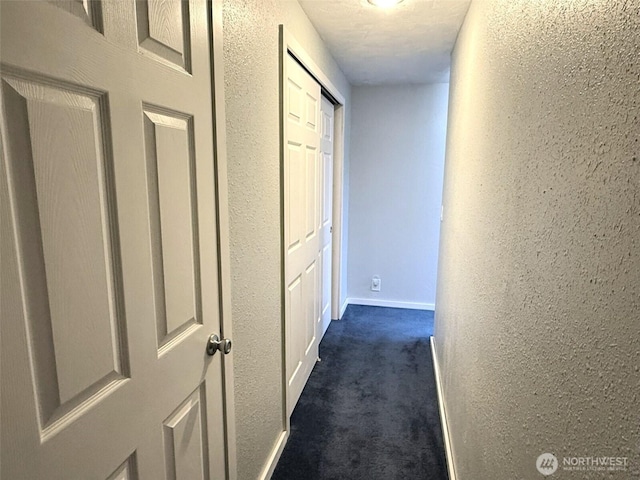 hall featuring dark carpet, a textured wall, and baseboards