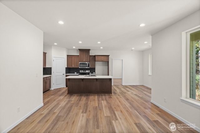 kitchen with light wood finished floors, tasteful backsplash, an island with sink, stainless steel appliances, and light countertops