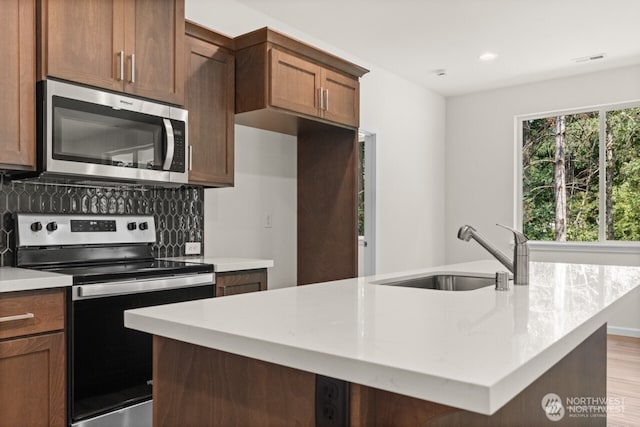 kitchen with light wood finished floors, a center island with sink, appliances with stainless steel finishes, a sink, and backsplash