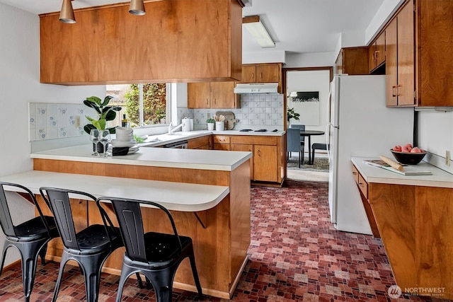 kitchen featuring backsplash, under cabinet range hood, light countertops, a peninsula, and brown cabinetry