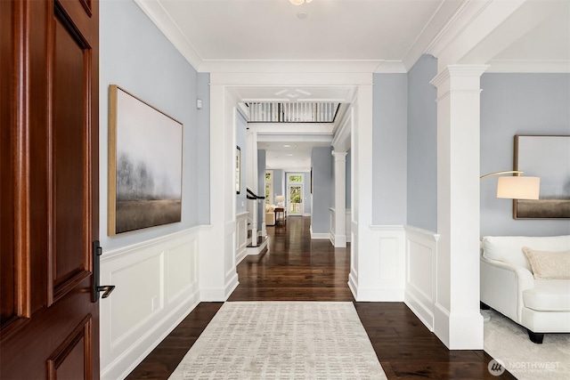 hallway featuring dark wood-style flooring, a decorative wall, ornamental molding, wainscoting, and ornate columns