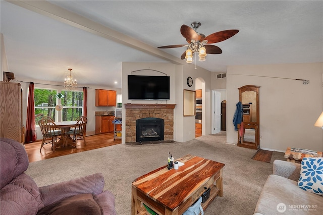 living room featuring visible vents, light colored carpet, vaulted ceiling with beams, a stone fireplace, and ceiling fan with notable chandelier