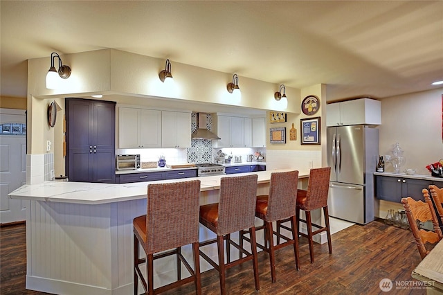 kitchen with a breakfast bar, wall chimney range hood, freestanding refrigerator, dark wood-style floors, and tasteful backsplash