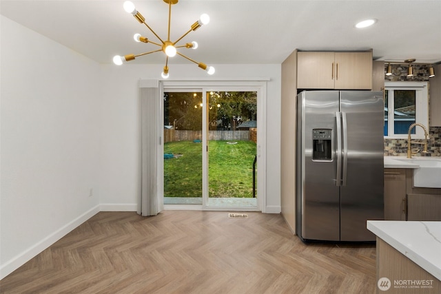 kitchen featuring stainless steel refrigerator with ice dispenser, tasteful backsplash, an inviting chandelier, a sink, and baseboards