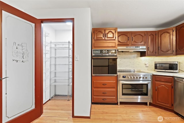 kitchen featuring under cabinet range hood, light countertops, appliances with stainless steel finishes, decorative backsplash, and light wood finished floors