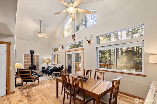 dining room with a ceiling fan, a wood stove, french doors, light wood-type flooring, and high vaulted ceiling