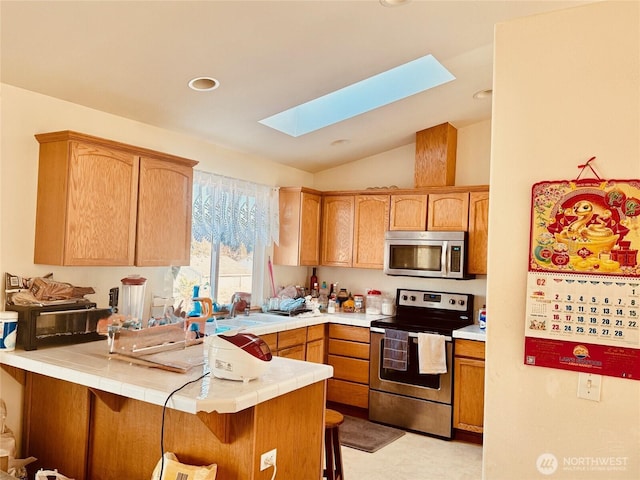 kitchen with tile counters, lofted ceiling with skylight, appliances with stainless steel finishes, a kitchen breakfast bar, and a peninsula