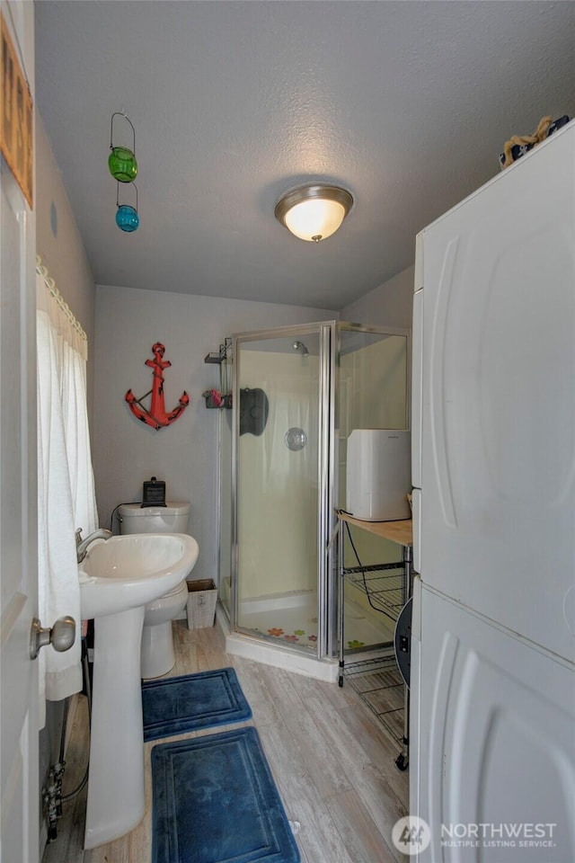 full bath featuring a textured ceiling, toilet, wood finished floors, stacked washer / drying machine, and a shower stall